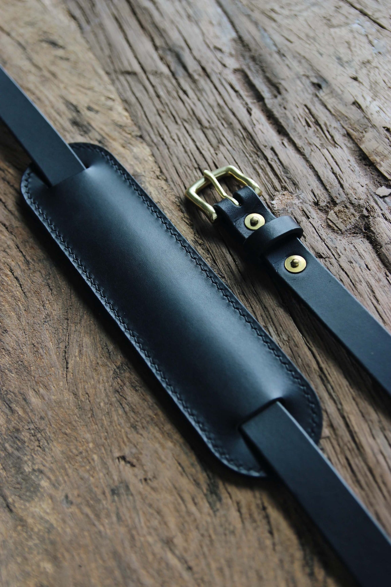 Detailaufnahme der Messingschnalle und des Schulterpads eines Leder Kameragurts in Schwarz auf einer Holzplatte.