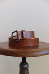 Ledergürtel für Herren in Braun zusammengerollt auf einem Stuhl mit einer Sitzfläche aus Holz.